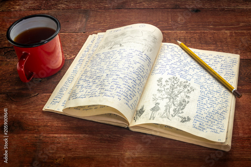 Slika na platnu Vintage expedition journal with a metal cup of hot tea on a rustic wood table  - handwriting and drawing in pencil