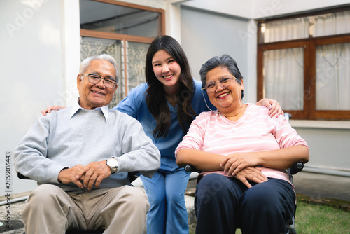 Asian happy elderly retired husband and wife with caregiver look camera on home garden, Older man in gray sweater sits beside Older woman wearing pink top while caregiver stands between them