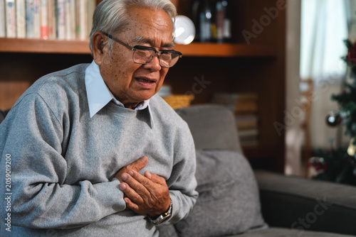 Senior asian man clutching chest having heart attack with worry. Elderly retired grandfather suffering from acute chest pain and face shows concern. old man breathlessness heart failure symptoms sofa