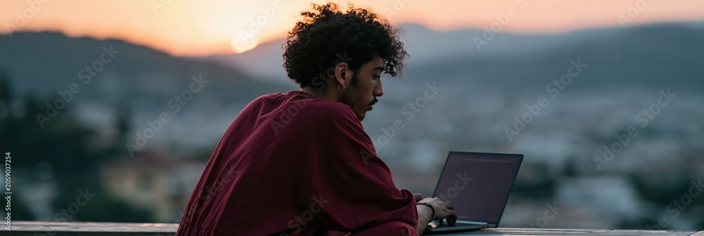 Fototapeta premium Man working on laptop outdoors at sunset on balcony with city view for remote work banner and productivity background scene