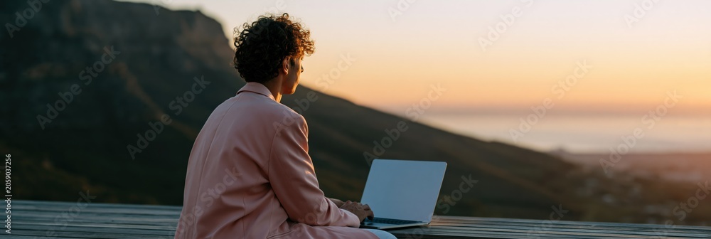 Fototapeta premium Young man on laptop at golden hour with mountain vista for digital nomad background and travel workspace wallpaper