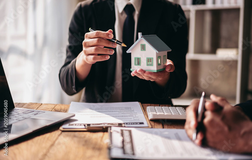Quadro su tela A man in a suit holding a model house in front of a desk with a laptop