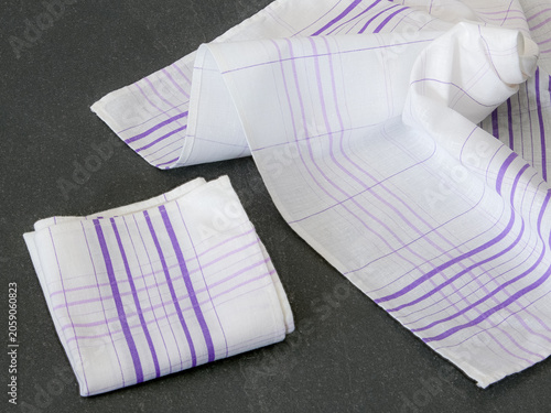 A neatly folded white cotton handkerchief with purple stripes and a similarly unfolded handkerchief on a textured dark background.