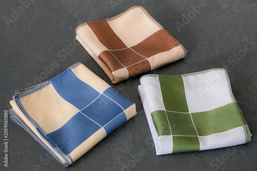 A set of folded cotton handkerchiefs featuring a minimalist cross pattern in blue, green, and brown on a textured dark background.