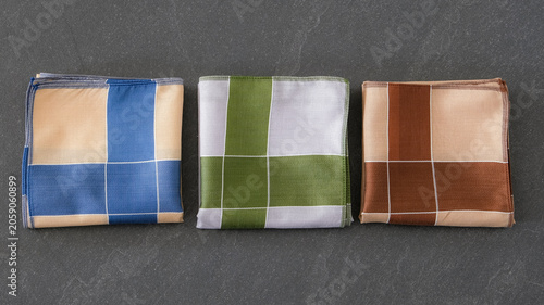 A set of folded cotton handkerchiefs featuring a minimalist cross pattern in blue, green, and brown on a textured dark background.