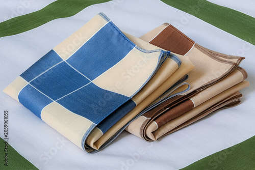 A blue-and-beige and a brown-and-beige folded handkerchiefs neatly arranged together on top of a green-bordered gray cotton handkerchief.