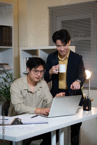 The collaborative work atmosphere between the two men in the office: the first, wearing a beige shirt, sits typing at a desk, while the other, in a dark suit, stands beside him.