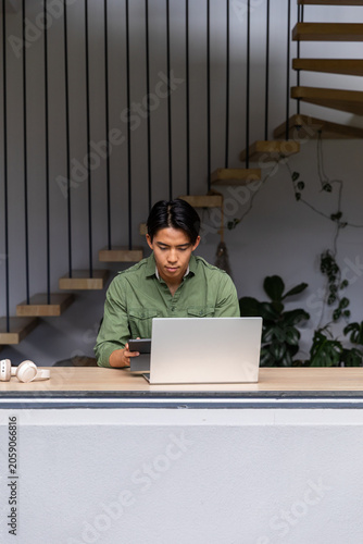 Asian man working in home workspace using silver laptop holding tablet with white headphones nearby