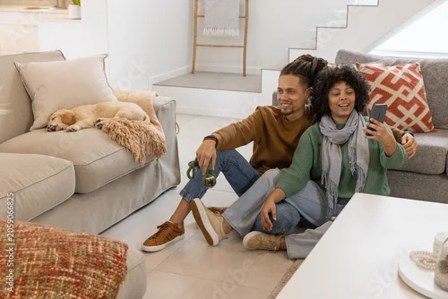 Fototapeta samoprzylepna African American couple sitting on tiled floor at home, holding smartphone, dog leash, sleeping dog