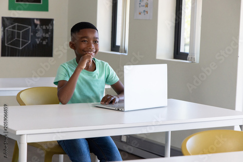 African American child sitting at white table using silver laptop on yellow chair wearing green tee