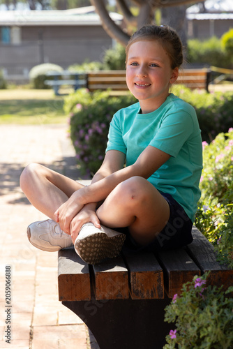 School-aged girl sitting cross-legged on wooden bench at park wearing teal T-shirt and white shoes