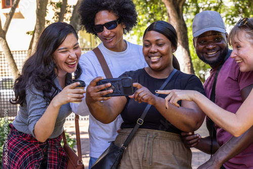 Diverse friends leaning in on paved path under trees, viewing horizontal smartphone and coffee cup