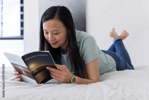 Asian mature woman lying on bed reading book ENDLESS SUNSETS in green tee and jeans