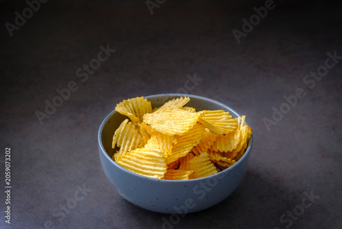 Golden, wavy chips fill a speckled bowl. Dark surface contrasts with crisp, inviting snack. Simple composition highlights snack's texture and color. Ideal for food, lifestyle, or casual eating scenes