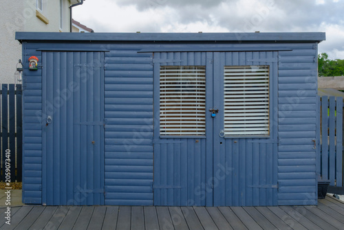 Blue wooden shed stands on a wooden deck. Windows with blinds shield the interior. Metal latch and door lock show secure access. Overcast sky adds calm, muted tone to scene. Ideal for garden, workshop