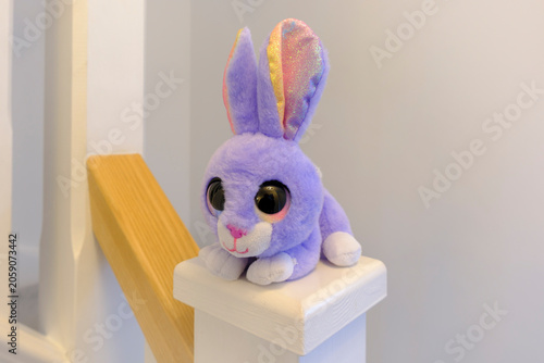 Purple bunny with sparkly ears sits atop white post. Soft plush toy rests near wooden staircase handrail. Large round eyes give sweet, innocent expression. Ideal for children's decor, bedtime stories