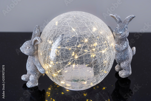 Glass sphere glows with warm fairy lights inside. Silver rabbits flank the sphere, posed protectively. Their textured bodies catch the soft illumination