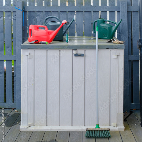 Garden tools rest neatly atop a storage unit. Bright red and green watering cans sit beside a broom. White storage box stands against a blue fence. Deck surface shows signs of recent rain