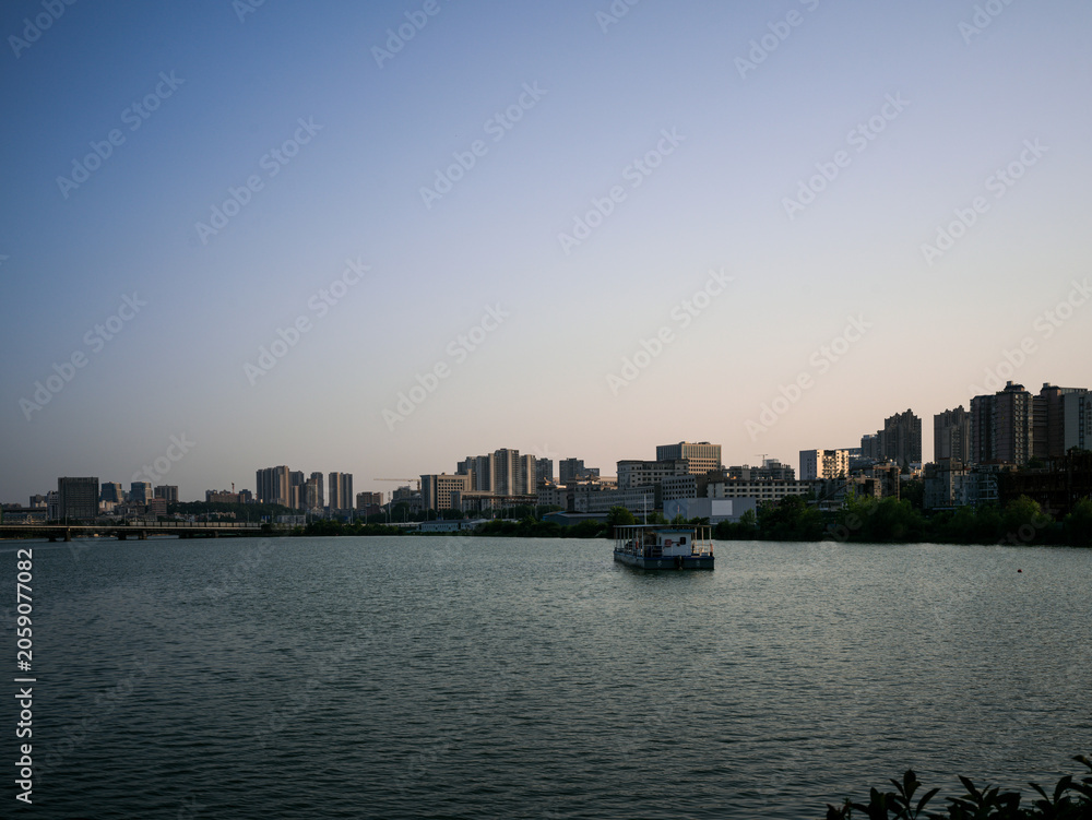 Fototapeta premium Small boat on calm lake water reflecting distant city skyline at evening twilight