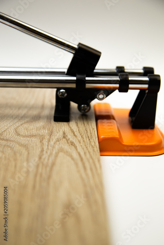 Close-up side view of an orange mechanical tile cutter, which fixed ceramic tiles of wooden colors, above it is a cutting disc on a white background. The concept of renovation, tiling, construction