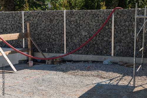 Construction site with a modern gabion stone fence and gravel-covered ground. A red flexible conduit runs across the scene, while wooden supports and scaffolding indicate ongoing building work