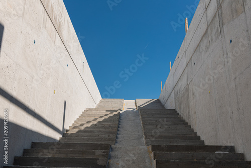 Concrete stairway under construction leads upward between tall reinforced concrete walls toward a clear blue sky. The minimalist architectural perspective highlights symmetry, geometric lines