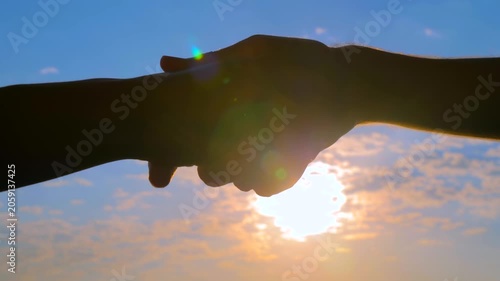 Silhouettes of man and woman are shaking hands against the sunrise or sunset sky - friendly handshake: close up view, sun lens flares. Greeting, agreement and friendship concept