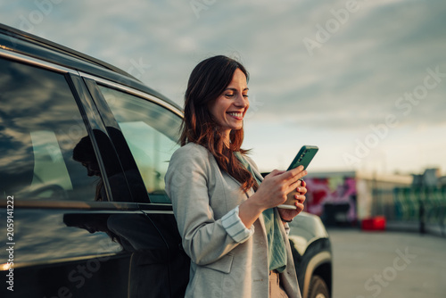 Foto Businesswoman standing by car, typing on her mobile phone, smiling while using m