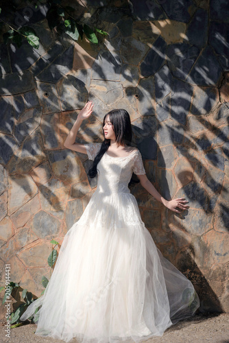 Young Asian Woman in White Dress Standing by Rustic Stone Wall with Natural Light