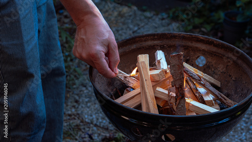 Tableau sur toile A man’s hand lights wooden kindling in a round metal barbecue grill using a match