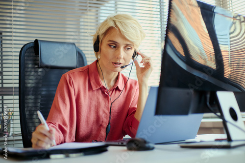 Obraz na plátne Medium shot of Caucasian middle aged businesswoman wearing headset, taking customer call and writing notes at desk