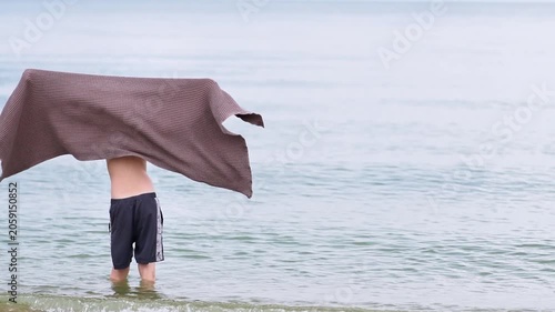 A teenager with a waffle towel in his hands basks in the sun under a light sea breeze on the seashore or ocean, peaceful tropical summer holiday, travel, relaxation, summer vacation,