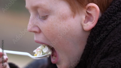 A red-haired teenager enjoys a piece of salad from a disposable utensil in a park on a cloudy day. A casual al fresco lunch and a moment in everyday life. High quality FullHD footage