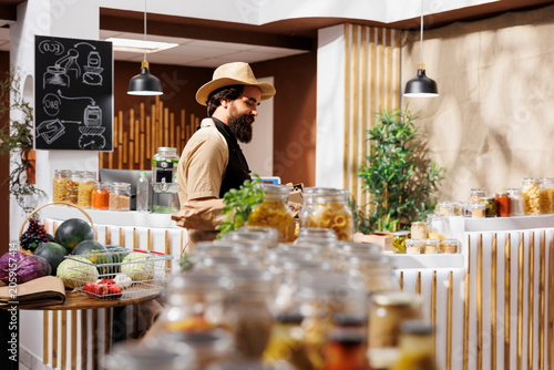Man in zero waste store filling shopping basket with healthy locally grown vegetables while environmentally responsible client buying organic non GMO food from local neighborhood shop.