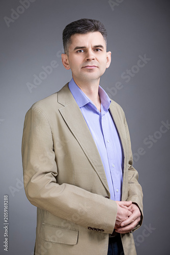 Portrait of Confident Positive Mature Gray-Haired Brunette Caucasian Man 50 Years Old Wearing Pale Jacket and Purple Violet Shirt Looking Straight With Closed Hands Over Gray