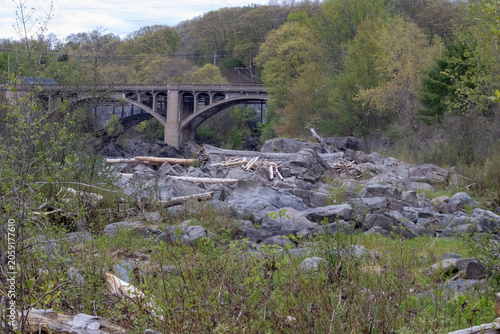 Fotografie Old concrete arch bridge spans a rocky riverbed strewn with driftwood logs, surr