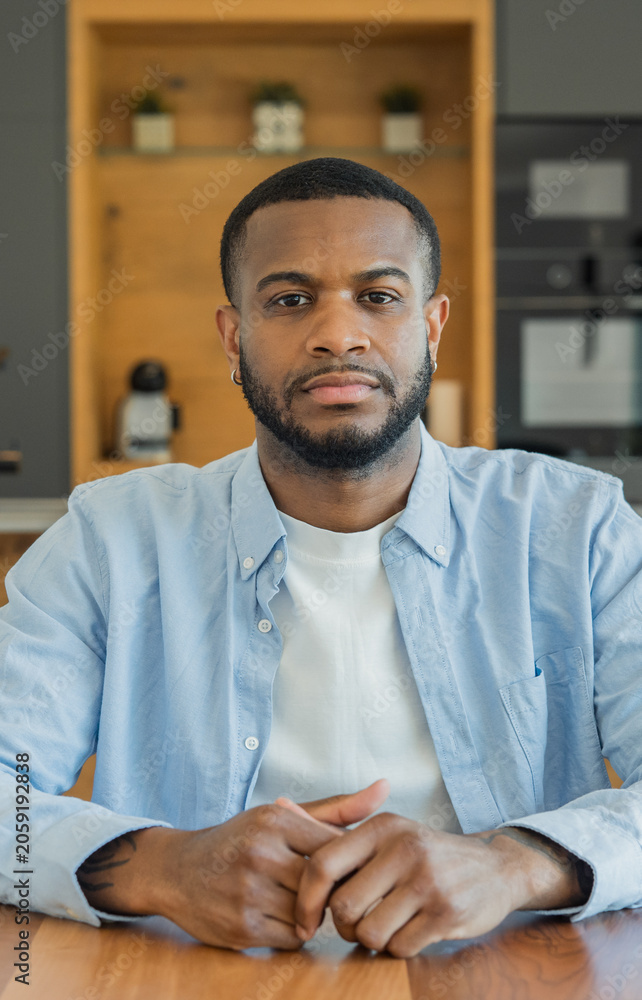 Fototapeta premium Young black entrepreneur sits at a wooden table in a relaxed home office, looking confidently at camera in a light blue shirt over white tee, embodying modern remote work