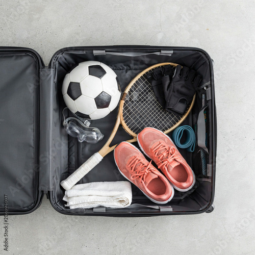 Suitcase contains a soccer ball, tennis racket, running shoes, towel, and goggles ready for a sports vacation trip.