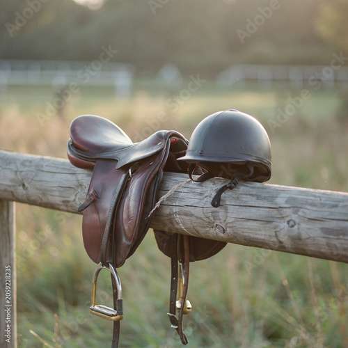 Horse saddle and a protective riding helmet rest balanced together upon a wooden fence rail at sunset outdoors.