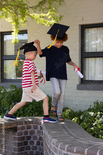 Two school-age boys wearing caps walking and balancing on curved brick wall at school with diplomas