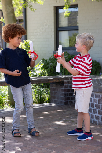 Diverse boys exchanging rolled white scrolls tied with red ribbons on paved brick patio near window
