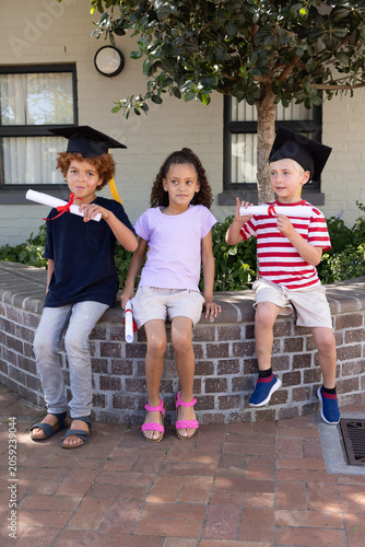 Three children sitting on low brick planter in courtyard wearing caps and holding red diplomas