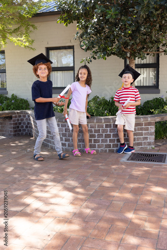 Diverse children wearing mortarboards holding red-ribboned diplomas on brick courtyard celebrating