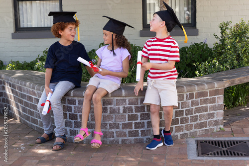 Three children sitting on curved planter at school wearing black caps, holding red-ribbon diplomas