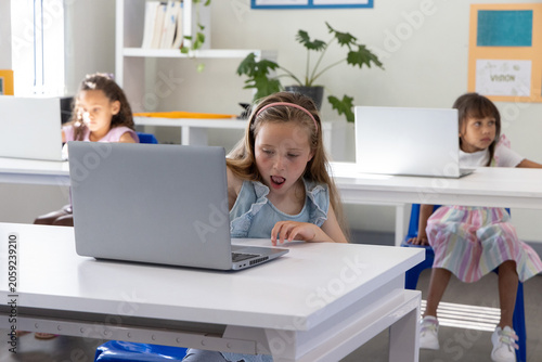 Фотография Three school-age girls using silver laptops at desks in classroom, center reacti