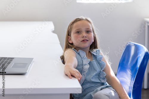 Female child sitting on blue chair at white table in study, wearing denim top, reaching laptop
