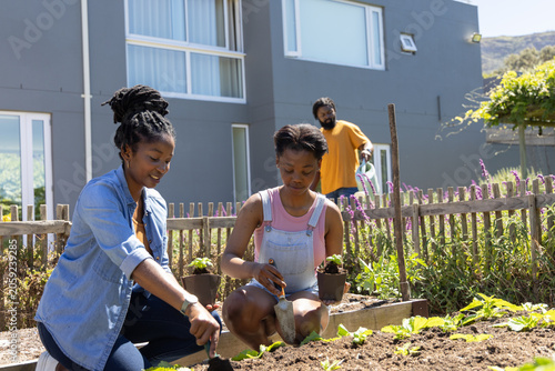 African family planting plants in backyard bed, teen daughter using trowel as dad holding green can