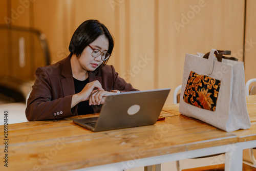 Fashionable Adult Asian Businesswoman sitting on a office desk wearing a glasses and brown suit working in laptop looking her wristwatch.