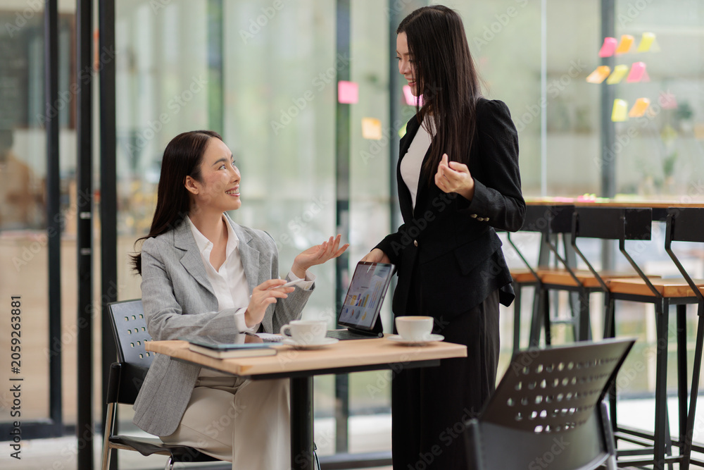 Fototapeta premium Two smiling asian businesswomen reviewing charts on a digital tablet during a collaborative meeting in a modern office, sharing ideas and analyzing data for strategy and growth
