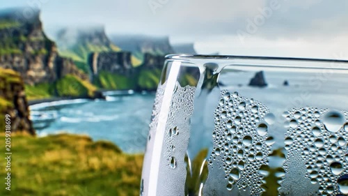 A close-up of a chilled glass with condensation droplets, overlooking a serene coastal landscape with rugged cliffs and calm waters